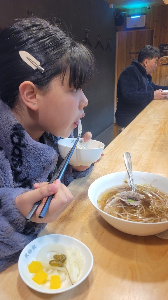 A bowl of rice noodles at a wooden counter in a Korean noodle shop, with pickled radish on the side